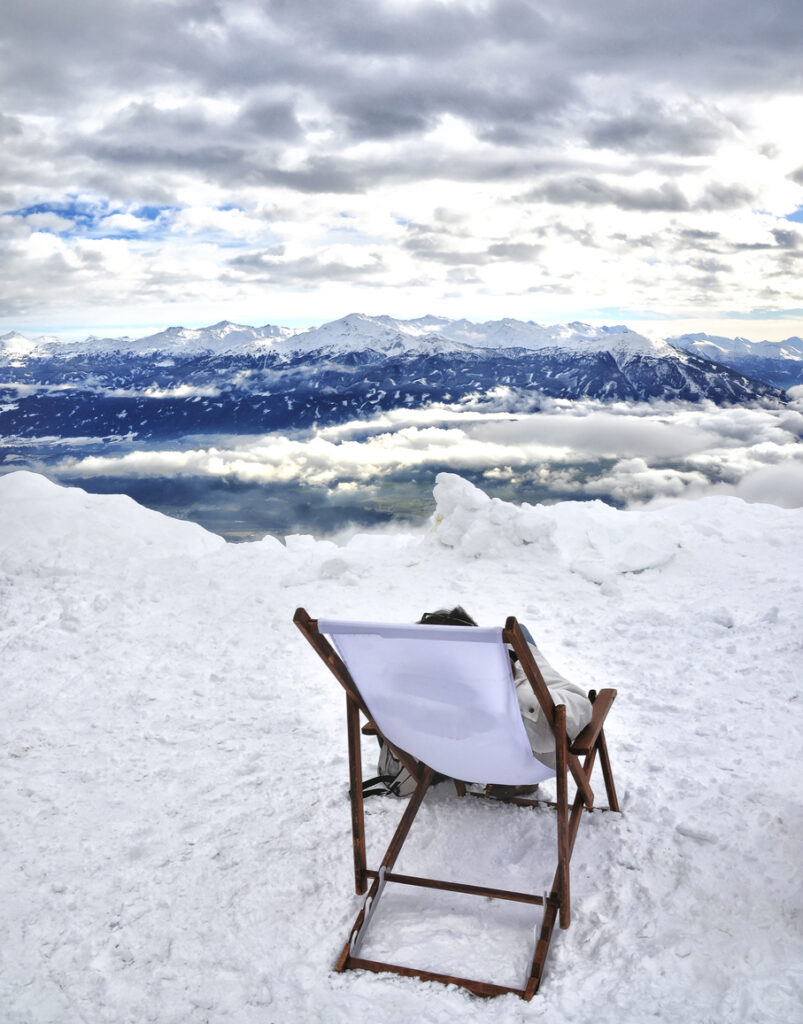 Person entspannt im Liegestuhl auf verschneitem Berggipfel mit Blick über das Inntal und die schneebedeckten Alpen bei Innsbruck
