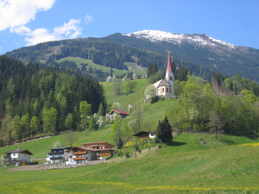 Traditionelles Tiroler Bergdorf mit weißer Kirche mit rotem Turm, Holzhäusern und grünen Wiesen vor bewaldeten Hängen und schneebedecktem Gipfel bei Fügen