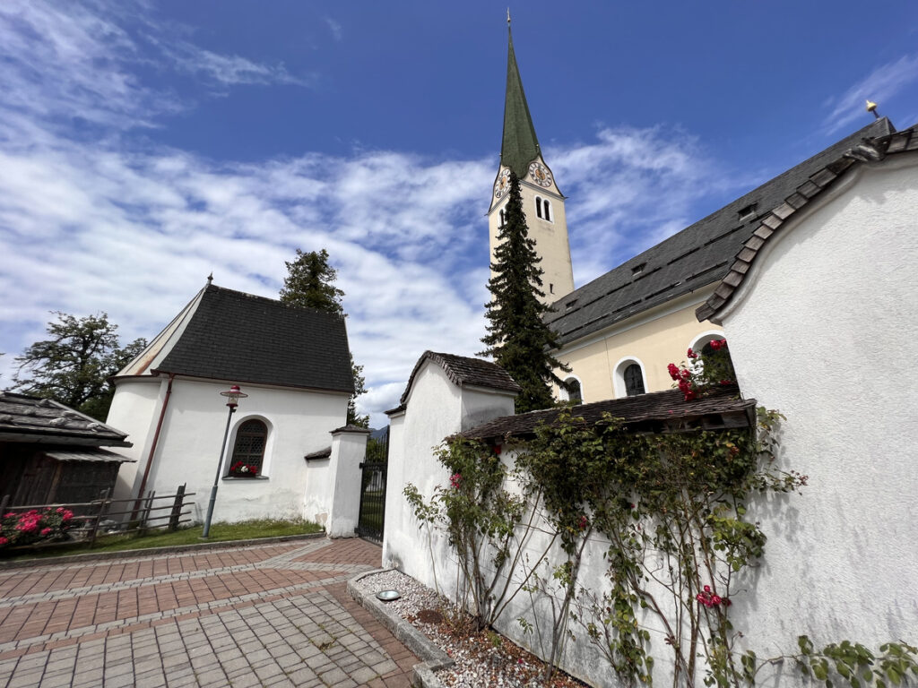 Historische Pfarrkirche mit hohem Kirchturm und weißer Kapelle im Ortszentrum von Kirchbichl unter blauem Himmel