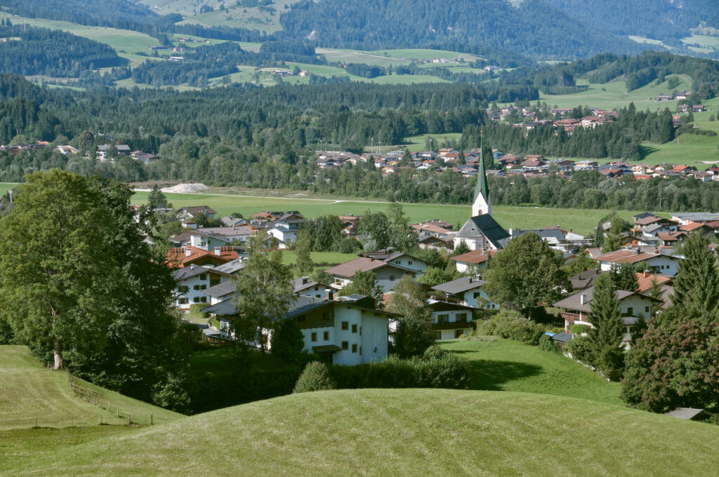 Panoramablick auf den Ort Kössen in Tirol mit Kirchturm, Wohnhäusern und grünen Wiesen vor bewaldeten Bergen