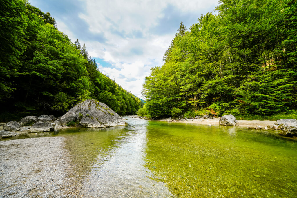 Klarer türkisgrüner Fluss mit Kiesufer und Felsen, umgeben von dichten grünen Wäldern in der Berglandschaft von Kramsach in Tirol